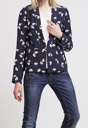 Žena má na sobě tmavě modrý květovaný blejzr přes bílý top, který je v kombinaci s tmavě modrými přiléhavými džíny, stojí s jednou rukou upravující blejzr.