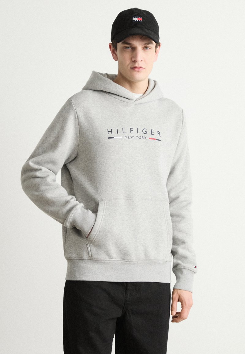 Siva pamučna dukserica s prednjim kengur đepom i rebrastim manžetama. Sadrži "HILFIGER NEW YORK" logo s crvenim i plavim detaljima. Crna kapa.