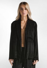Blazer in velluto a costo nero con una finitura testurizzata, con ampie rever e maniche lunghe, indossato sopra un top a coste marrone. Linee pulite e vestibilità rilassata.