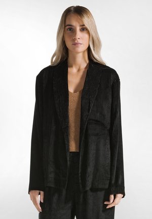 Zwarte corduroy blazer met een gestructureerde afwerking, voorzien van brede revers en lange mouwen, over een bruin geribbeld topje. Strakke lijnen en een ontspannen pasvorm.