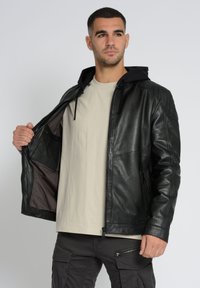 Schwarze Lederjacke mit Kapuze, vorderem Reißverschluss und gesteppten Schulterdetails. Über einem hellbeigen Hemd getragen, das Innentaschen zeigt.