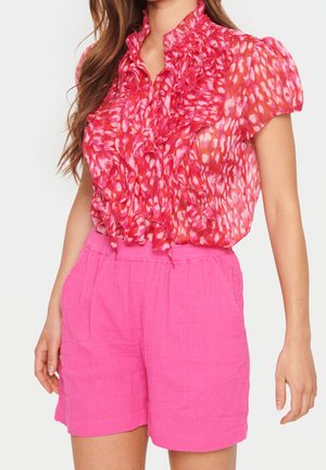 Lyserød bluse med korte ærmer, flæsedetalje og blomstermønster, kombineret med højtaljede lyserøde shorts i et tekstureret stof.