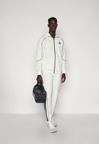 Fato de treino atlético branco com detalhes e riscas pretas. Modelo segurando uma mochila desportiva preta com logótipo branco. Tênis são cinza claro.