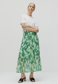 Grøn floral maxi-nederdel med en tiered kant, der har en gennemsigtig struktur og mønstre af blade og blomster, stylet med en hvid t-shirt og sorte sandaler.