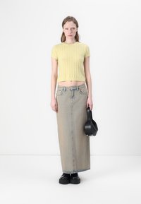 Top scurt tricotat de un galben deschis, cu mânecă scurtă și model cu cabluri, asortat cu o fustă lungă din denim decolorat. Modelul ține o geantă de mână neagră.