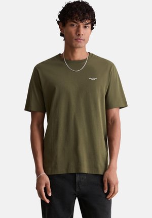 Junger Mann mit lockigem Haar, der ein olivgrünes T-Shirt mit kleinem weißem Text und schwarze Hose trägt, steht vor einem einfarbigen Hintergrund.
