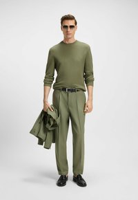 Olive green outfit featuring a textured knit sweater, tailored trousers, and a matching coat draped over the arm; sleek black shoes and sunglasses.