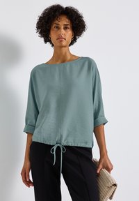 Blusa verde chiaro, dal fit morbido, con maniche a tre quarti, scollatura a girocollo e coulisse all'orlo, superficie testurizzata. Abbinata a pantaloni neri.