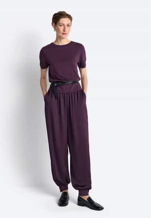 Vrouw staat met handen in de zakken, draagt een paarse top met korte mouwen en bijpassende losse broek met een zwarte riem en zwarte schoenen.