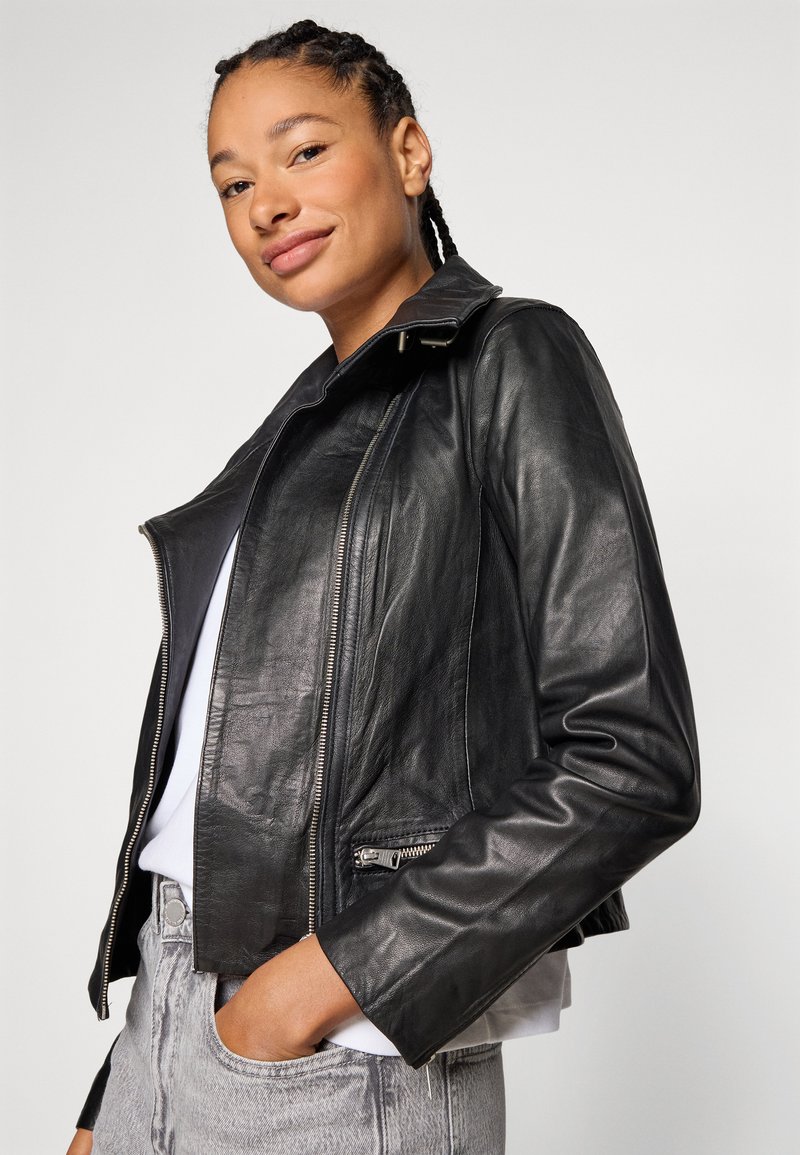 Schwarze Leder-Bikerjacke mit taillierter Passform, Reißverschlusstaschen und Druckknopfkragen, kombiniert mit einem weißen Oberteil und grauen Jeans.