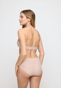 Donna di spalle che indossa un reggiseno a fascia beige chiaro e biancheria intima a vita alta, con due piccoli tatuaggi visibili sulle braccia.