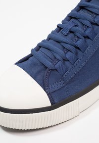 Baskets bleu marine présentant un dessus en tissu texturé, une capuche en caoutchouc blanc classique et une semelle noire avec des détails nervurés.