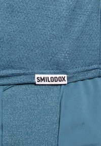 Turkusowy top sportowy z fakturowanej tkaniny, ze gładką sekcją i czarną metką "SMILODOX" wszytą na dole.