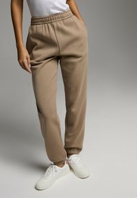 Beżowe dresy z elastycznym paskiem w talii, bocznymi kieszeniami i ściągaczami na nogawkach. W zestawie z białymi sneakerami z delikatnym różowym akcentem.