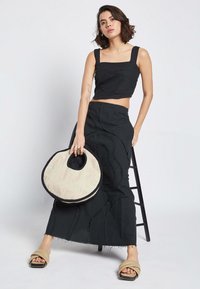 Črn usnjeni top s kvadratnim izrezom in širokimi naramnicami, v kombinaciji s črno dolgo krilo s teksturiranimi vzorci. Drži okroglo pleteno torbo.