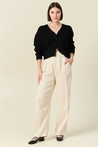 Cardigan noir en tricot avec un décolleté en V profond, superposé sur un pantalon large crème. Le modèle porte des chaussures plates noires. Design et texture simples.