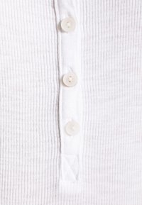 Vit ribbad henley-topp med tre små, runda, beige knappar längs mitten av knappslån och en triangulär detalj nära nederkanten.