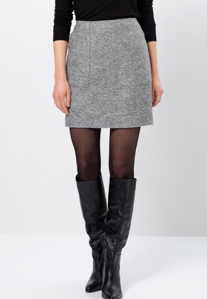 Femme portant une jupe en laine grise, des collants noirs transparents, des bottes en cuir noires montantes jusqu'aux genoux et un haut noir à manches longues sur un fond uni.