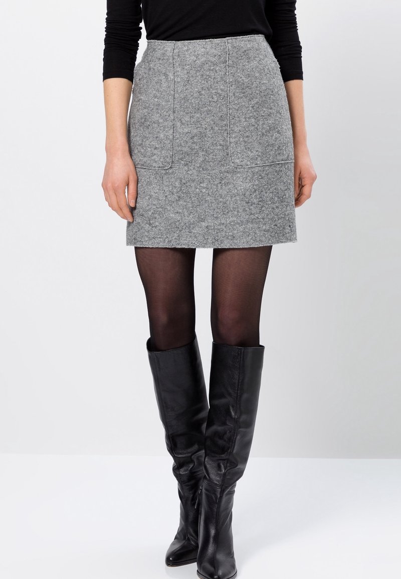 Femme portant une jupe en laine grise, des collants noirs transparents, des bottes en cuir noires montantes jusqu'aux genoux et un haut noir à manches longues sur un fond uni.