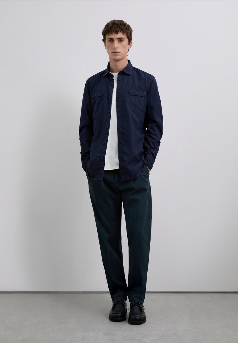 Marineblaue Hemdjacke mit zwei Brusttaschen, kombiniert mit dunkelgrünen Hosen und schwarzen Schuhen. Schlichtes Design und taillierte Silhouette.