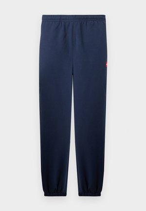 Pantalon de survêtement bleu marine longueur totale avec taille et chevilles élastiques, comprenant une petite étiquette de logo rouge sur la hanche droite.