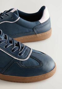 Navy blue sneakers with a smooth leather upper, white accents, and a textured heel. Features a rubber outsole with a brown finish and lace closure.