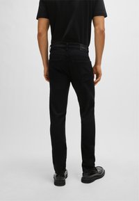 Black denim jeans with a slim fit, rear pockets, and a subtle brand label patch on the waistband. Smooth texture and tapered design.