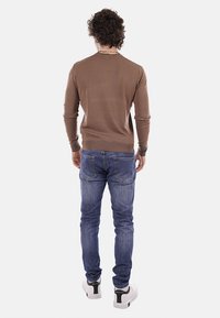 Homme aux cheveux bouclés portant une chemise marron à manches longues, un jean bleu et des baskets blanches, debout de dos sur un fond uni.