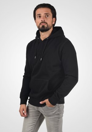 Sudadera negra de tela suave, con un bolsillo tipo canguro, capucha con cordón y puños acanalados. Combinada con pantalones gris claro.