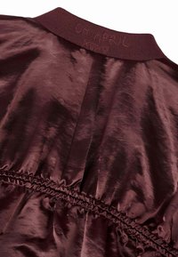 Burgundinė satino suknelė su gūžtuota apykakle, susiraukšlėjusiu juosmeniu ir subtiliu blizgesiu bei lygiu paviršiumi. Ant apykaklės yra išspaustas užrašas "04 BALANDIS".