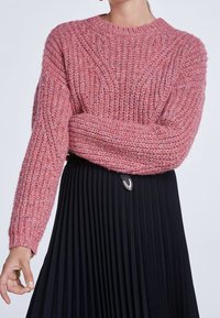 Rosa gestrickter Pullover mit geribbelter Textur und langen Ärmeln, kombiniert mit einem schwarzen Faltenrock. Auffällige Details sind ein runder Ausschnitt und sichtbare Nähte.