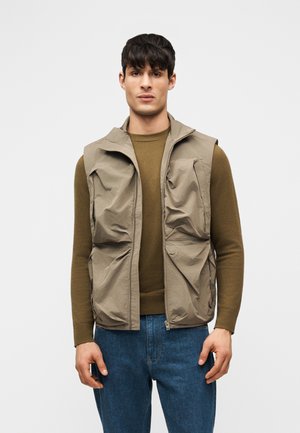 Mand iført olivengrøn langærmet skjorte, beige ærmeløs vest med lynlås og store lommer samt blå jeans mod en ensfarvet baggrund.