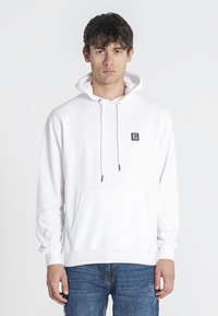 Sudadera blanca de tejido suave, con un bolsillo frontal, capucha con cordón y un pequeño logo negro en el pecho. Llevada con jeans azules.