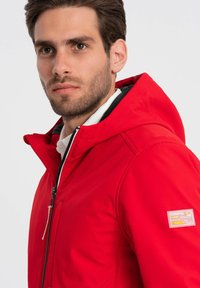 Rød zip-up jakke med hætte, der har en frontlomme, kontrast sort foer og et mærke på ærmet. Glat, vandafvisende stof.