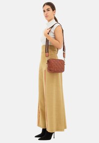 Borsa a tracolla in pelle marrone trapuntata con superficie testurizzata e tracolla a motivo contrastante. Il modello indossa pantaloni beige a vita alta e tacchi neri.