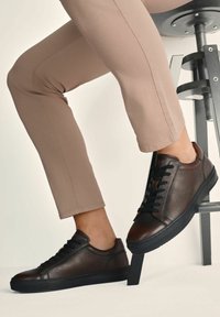 Nogi w beżowych spodniach, w ciemno-brązowych skórzanych sneakersach z czarnymi podeszwami, jedna stopa oparta o nogę czarnego stołka na białym tle.