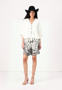 Hvit oversized topp med knyting foran, sammen med mønstrede shorts med svart broderi. Modellen har på seg en svart bredbremmet hatt og beige hæler.