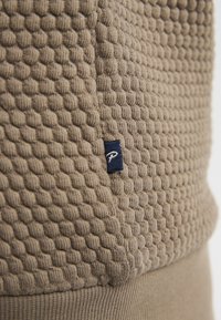 Texturerad beige tyg med ett bikake-mönster; innehåller en liten blå etikett med en vit logotyp. Slät nedre del.
