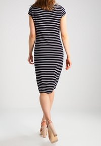 Robe midi rayée bleu marine et blanche avec des manches courtes, silhouette ajustée et détails de fronces sur les côtés. Portée avec des sandales à talons carrés couleur nude.