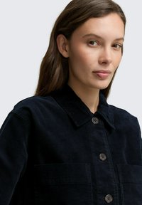 Jonge vrouw met lang bruin haar, gekleed in een donkermarine corduroy jack met knopen, kijkt iets naar de zijkant tegen een effen achtergrond.