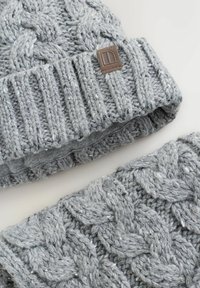 Grå stickad mössa och matchande halsvärmare, med ett kabelstickat mönster. Mössan har en ribbad kant och en liten brun logotyp-patch.