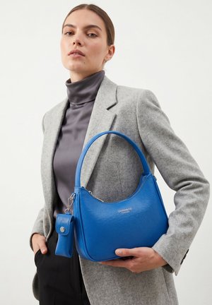 Femme portant un blazer gris et un col roulé sombre tient un sac à main en cuir bleu vif avec une petite pochette attachée.