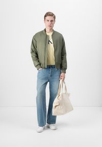 Olivengrønn bomberjakke, gul t-skjorte med logo, lyseblå vide bukser, hvite sneakers, holder en beige tote bag med logodetaljer.