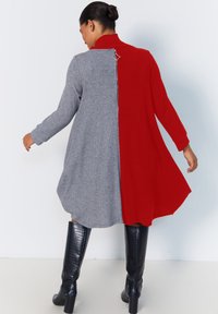 Robe en tricot grise et rouge avec un col haut, une coupe asymétrique et un zip dorsal prominent. Présente des manches longues et une silhouette fluide.
