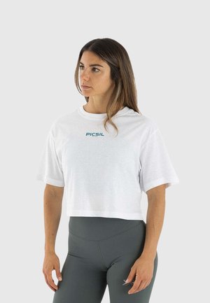 Vrouw met lang bruin haar, gekleed in een wit cropped T-shirt en donkere hooggetailleerde leggings, kijkt naar links tegen een effen achtergrond.