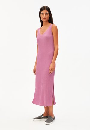 Femme debout portant une robe midi rose côtelée sans manches avec des fentes latérales et des chaussures décontractées grises à enfiler sur fond blanc.