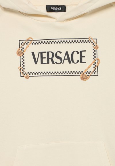 Hoodie de cor creme com um logótipo preto "VERSACE" emoldurado por um padrão de xadrez e adornado com alfinetes de segurança dourados.