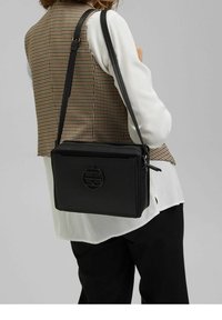 Donna con un gilet a scacchi marroni, una camicia bianca e pantaloni neri, che porta una borsa a tracolla in pelle nera con un logo circolare.
