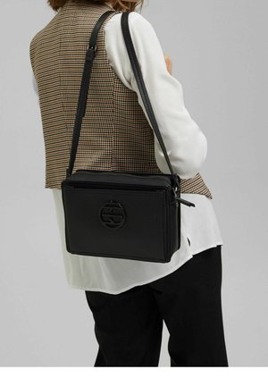 Kvindelig iført en brunternet vest, hvid bluse og sorte bukser, bærende en sort læder crossbody taske med et rundt logo.