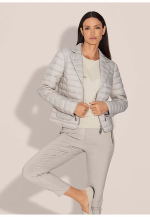 Frau trägt eine hellgraue gesteppte Jacke, passende Hose und weiße Schuhe und posiert mit einem angehobenen Bein vor einem einfarbigen beigen Hintergrund.